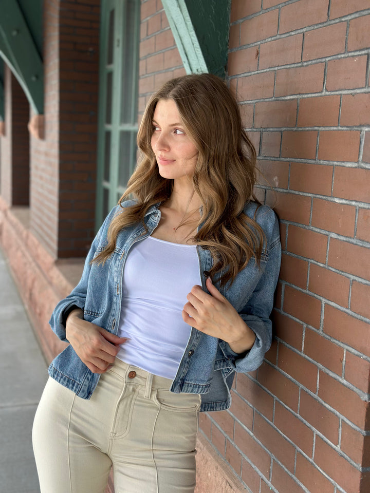 A woman with long brown hair leans against a red brick wall, wearing a light blue denim jacket over a white tank top and beige pants.