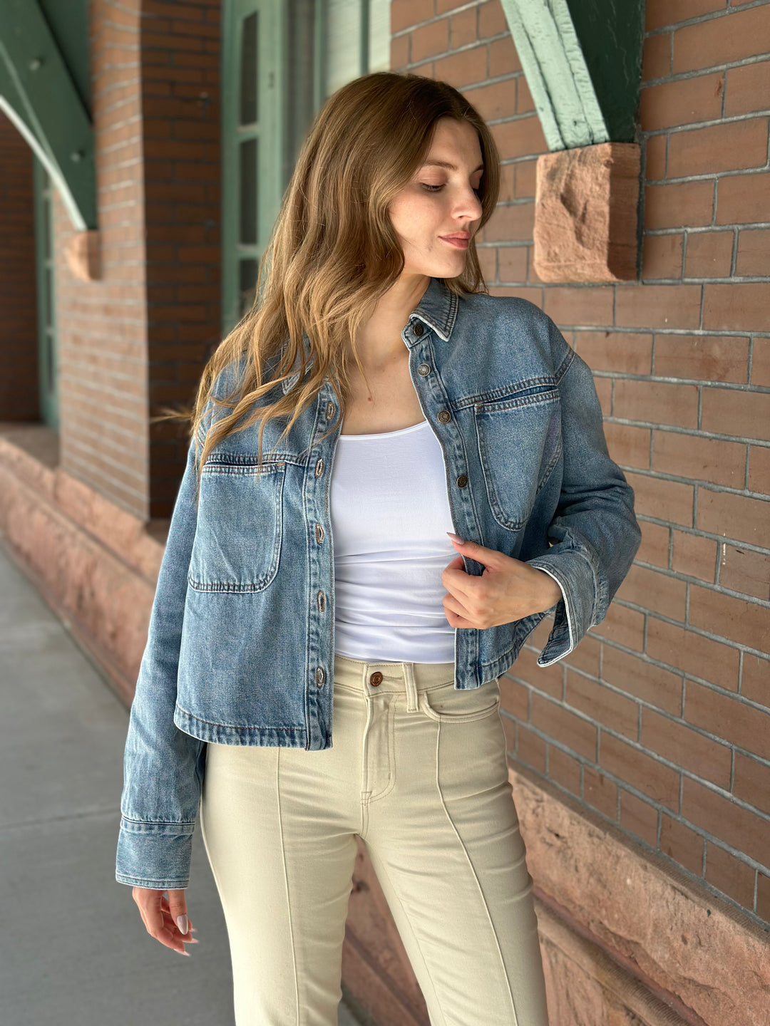 A woman wearing a light-wash blue denim jacket over a white tank top and beige pants, standing outdoors against a brick wall.