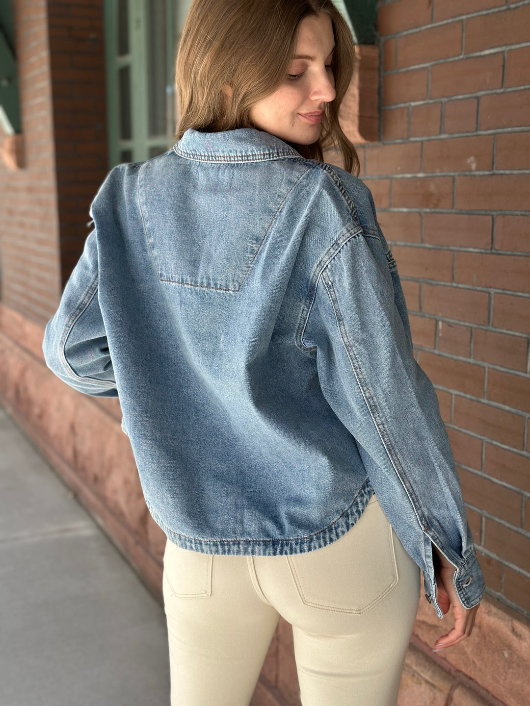 A woman wearing a light blue denim jacket and cream pants, photographed from behind in an outdoor urban setting with a brick wall background.