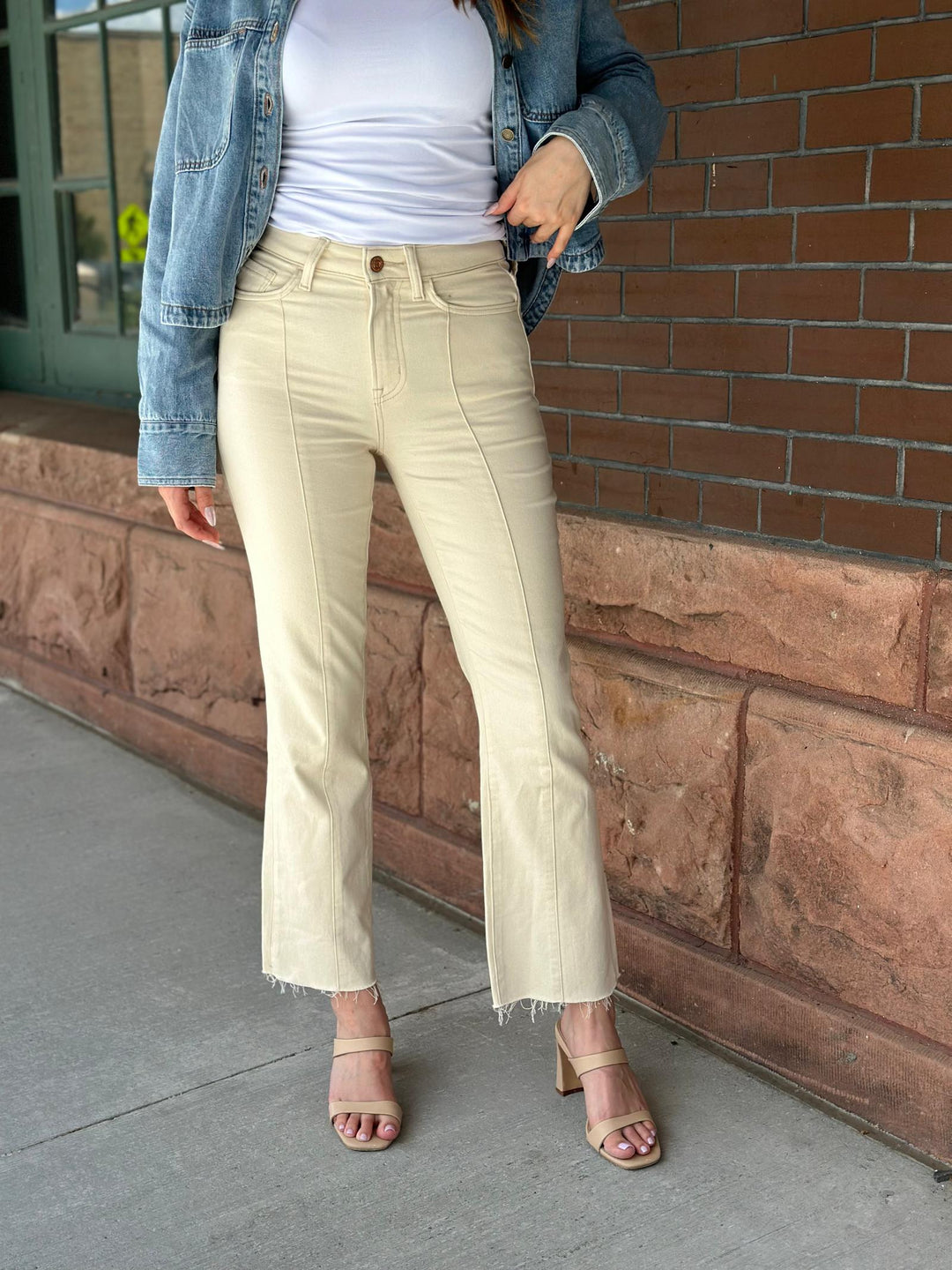 A person wearing cream-colored cropped flare jeans with a frayed hem, styled with a white top, a blue denim jacket, and beige heeled sandals, standing outdoors against a brick wall.