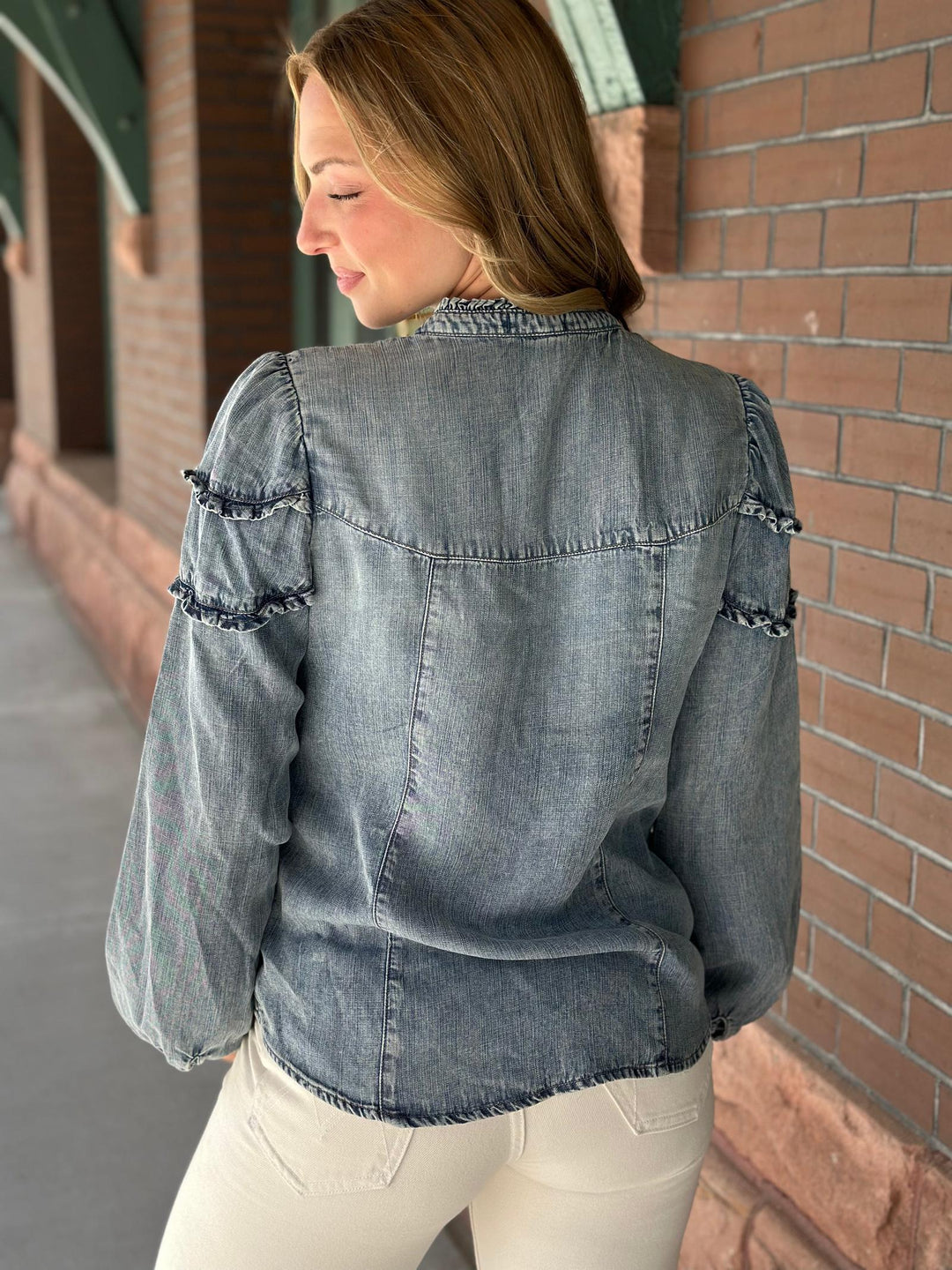 A woman wearing a faded blue-gray denim jacket with ruffled sleeve details, shown from the back in an outdoor setting next to a brick wall.