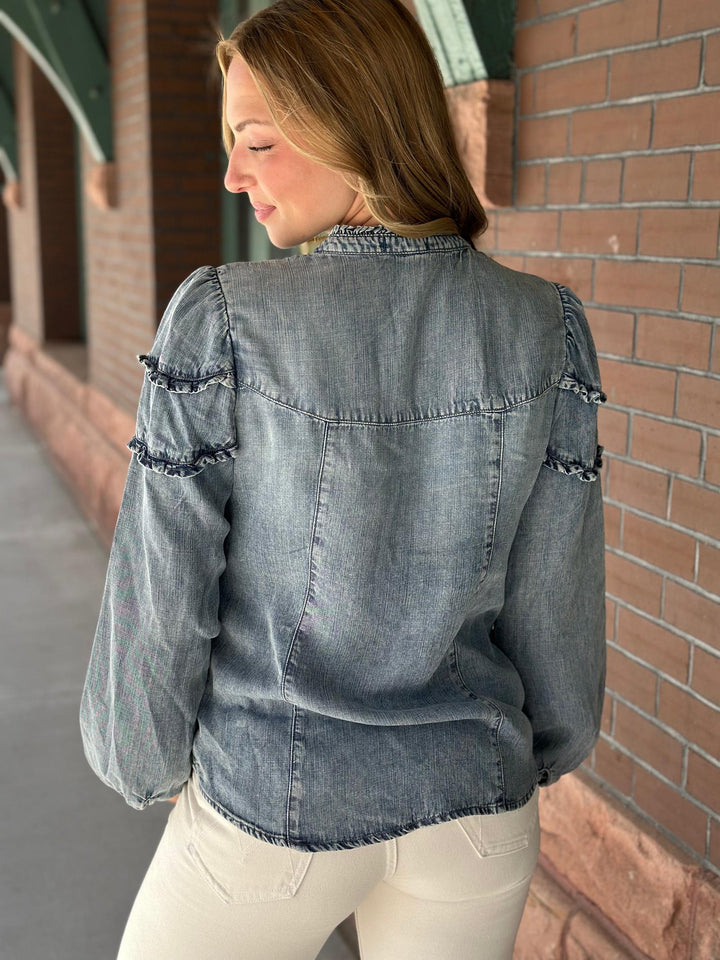 A woman wearing a faded blue-gray denim jacket with ruffled sleeve details, shown from the back in an outdoor setting next to a brick wall.