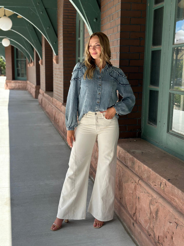 A woman stands on a covered outdoor walkway wearing a light-wash blue denim shirt with ruffle details and off-white wide-leg pants.