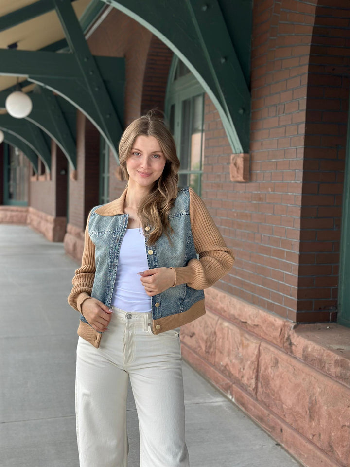 A woman stands outdoors on a walkway in front of a brick building with green metal arches, wearing a light blue denim jacket with tan ribbed knit sleeves and collar, a white tank top, and off-white pants.