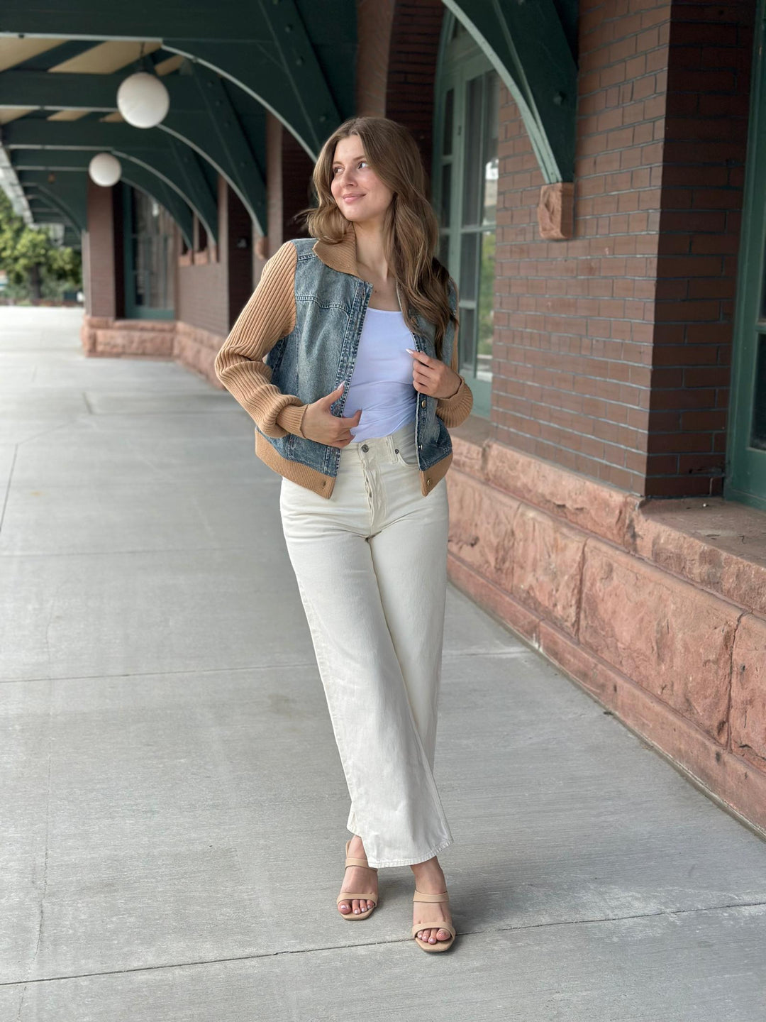 A woman stands on a sidewalk in front of a brick building with green trim, wearing a tan ribbed knit cardigan, a denim vest, a light blue tank top, off-white cropped pants, and beige heeled sandals.