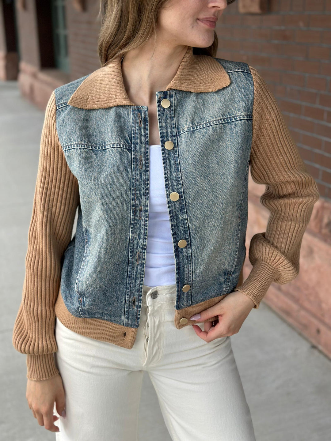 A woman wearing a cropped denim jacket with tan ribbed knit sleeves and collar, paired with a white top and white pants, standing outdoors against a brick wall.