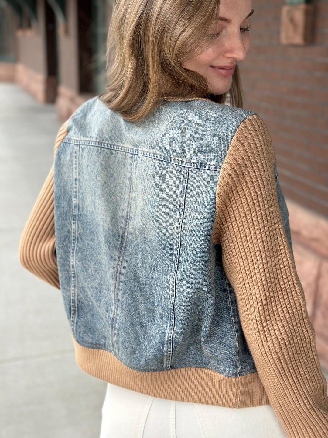 A woman shown from the back wearing a light blue denim vest with tan ribbed knit sleeves and hem, photographed outdoors on a sidewalk.