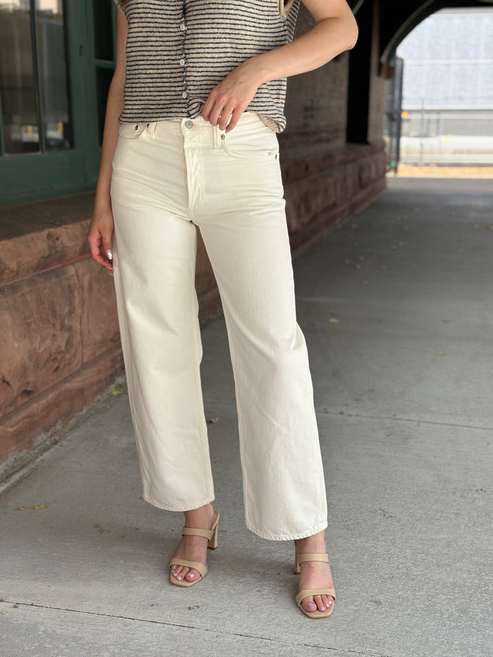 A person wearing off-white wide-leg cropped pants, a striped sleeveless top, and beige heeled sandals, standing on a concrete walkway under a covered passageway.