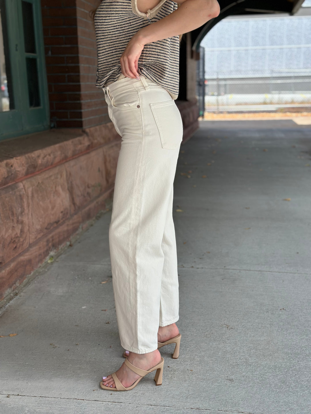 A person wearing off-white wide-leg jeans with side slits, paired with a striped top and beige heeled sandals, standing on a concrete walkway next to a brick building.