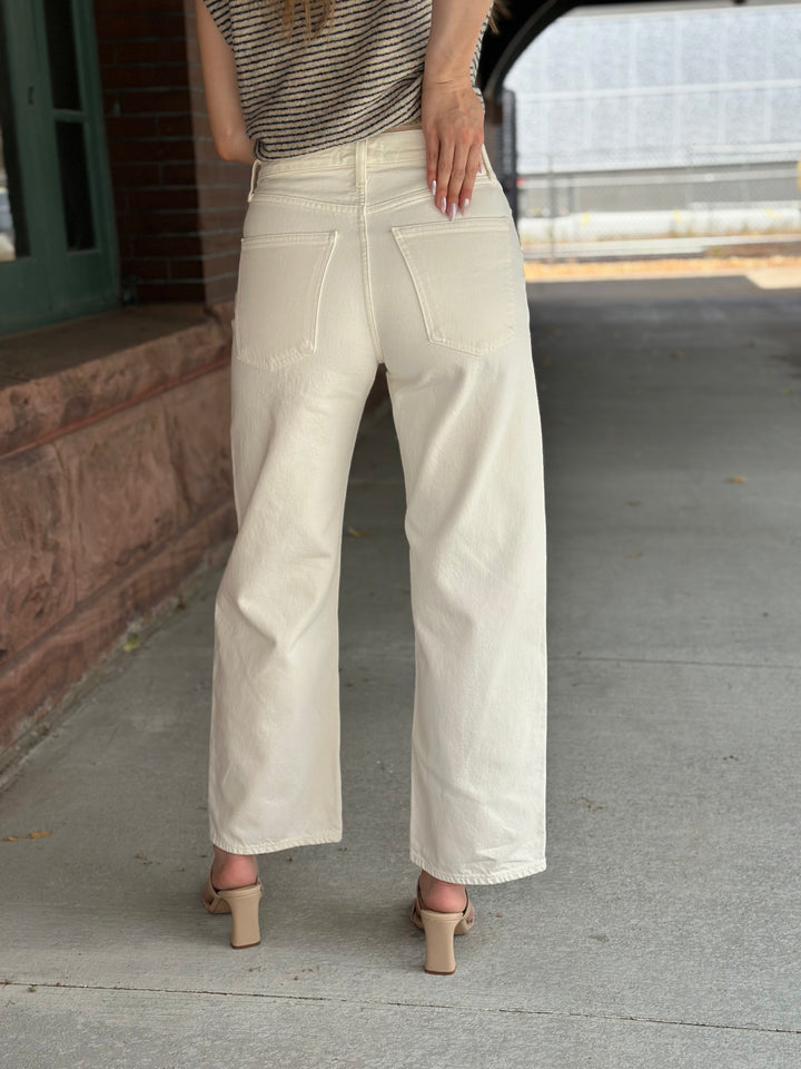 A person shown from the back wearing off-white wide-leg jeans, a striped top, and beige block-heel sandals, standing on a concrete sidewalk.