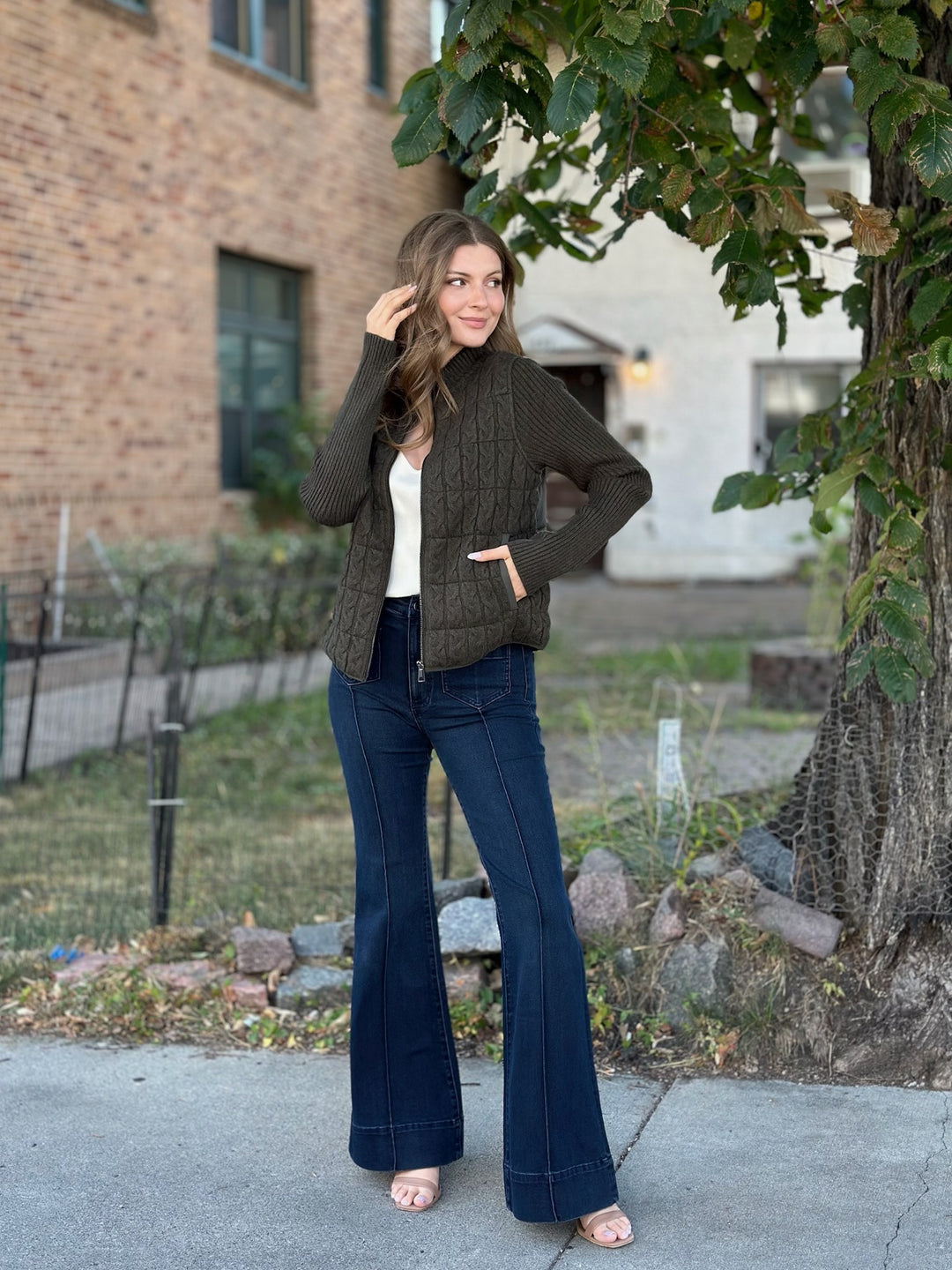 A woman stands outdoors on a sidewalk wearing a dark olive green cable-knit zip-front cardigan over a white top, paired with dark blue flared jeans and pink sandals. A brick building, grass, and a tree are visible in the background.