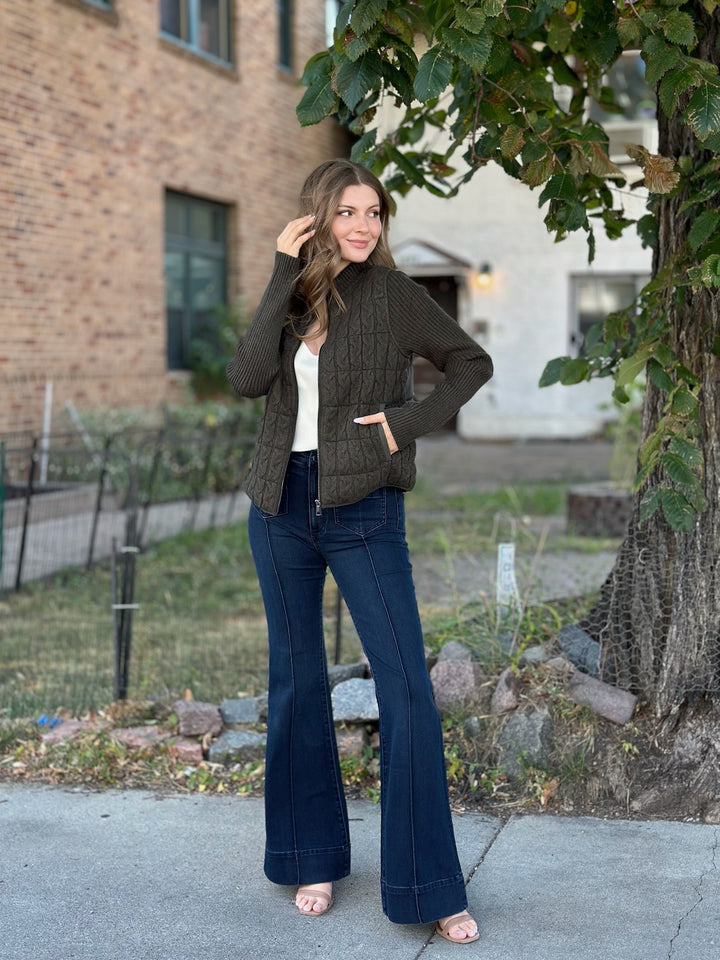 A woman stands outdoors on a sidewalk wearing a dark olive green cable-knit zip-front cardigan over a white top, paired with dark blue flared jeans and pink sandals. A brick building, grass, and a tree are visible in the background.