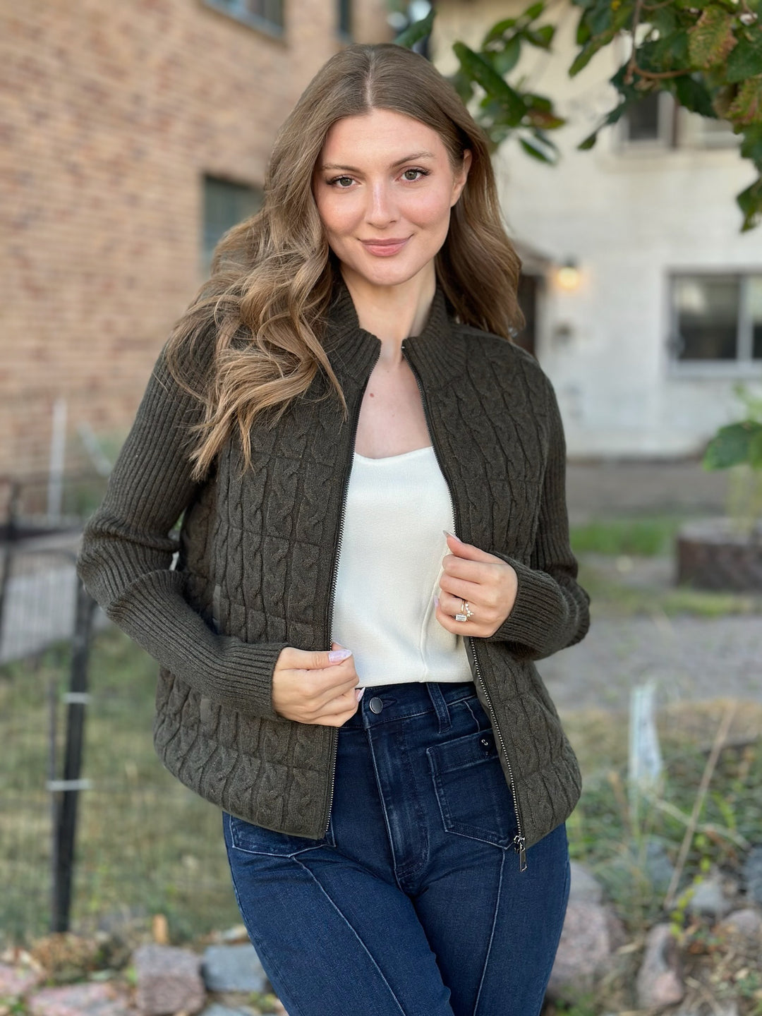 A woman wearing a dark olive green cable-knit zip-up jacket over a white top and blue jeans, standing outdoors in front of a brick building.