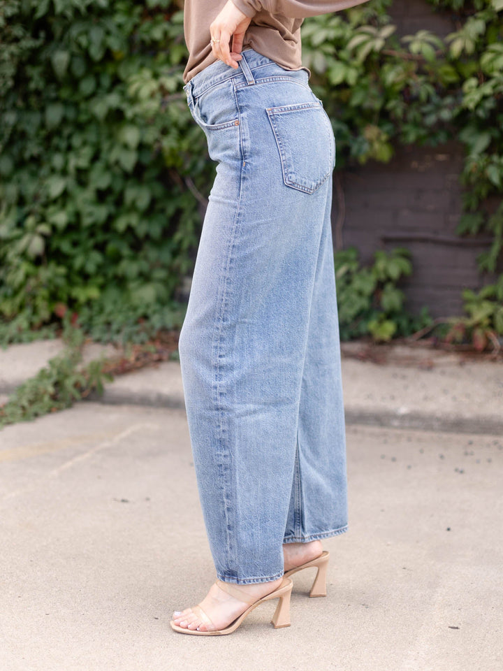 A person wearing light-wash wide-leg denim jeans, a tan top, and beige heeled sandals, standing outdoors on a paved surface with greenery and a dark wall in the background.