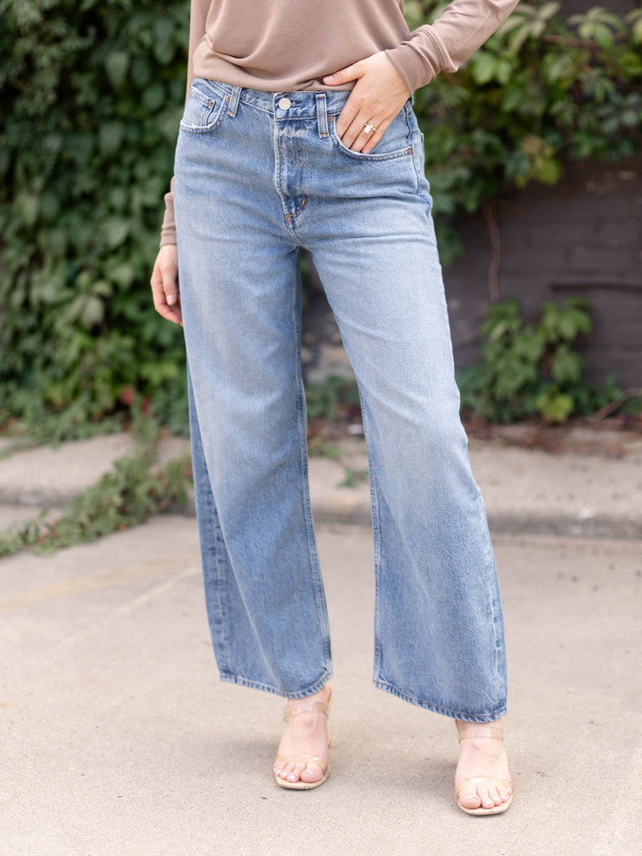 A person wearing light-wash blue wide-leg jeans, a taupe long-sleeve top, and clear high-heel sandals, standing outdoors on a paved surface with greenery in the background.