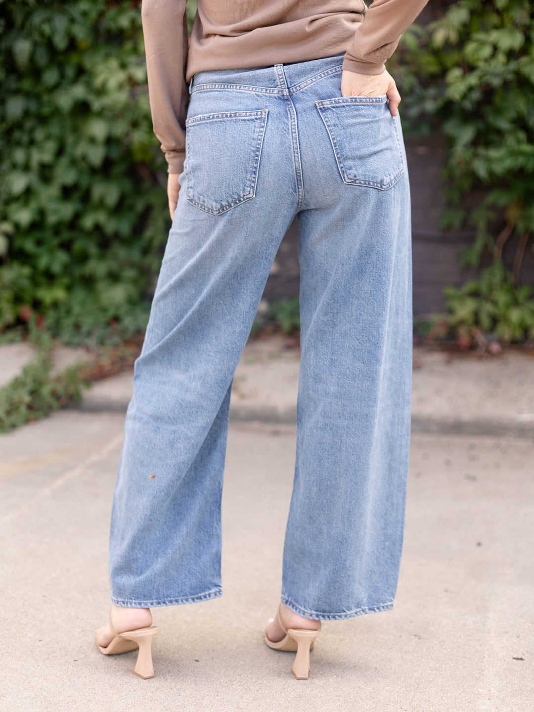 A person shown from the back wearing light-wash wide-leg denim jeans, a tan long-sleeve top, and beige heeled sandals, standing outdoors on a paved surface with greenery in the background.