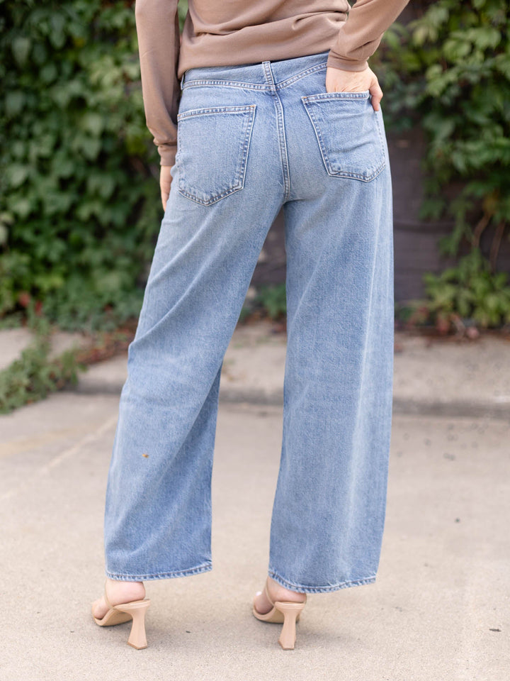A person shown from the back wearing light-wash wide-leg denim jeans, a tan long-sleeve top, and beige heeled sandals, standing outdoors on a paved surface with greenery in the background.