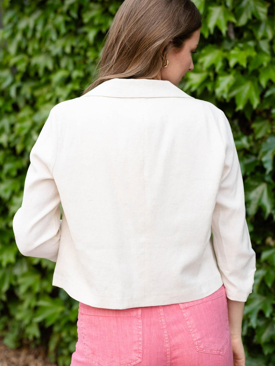 Woman from behind wearing a natural linen blend cropped 3/4 sleeve open blazer over pink pants.