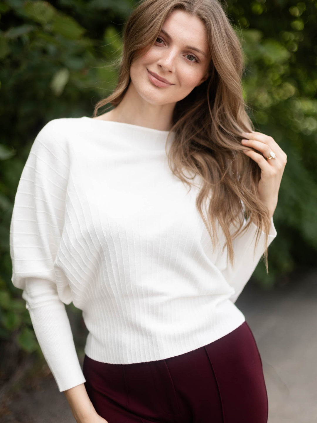 A woman wearing a white ribbed knit sweater with long balloon sleeves and a boat neckline, paired with burgundy pants, standing outdoors against a blurred green foliage background.