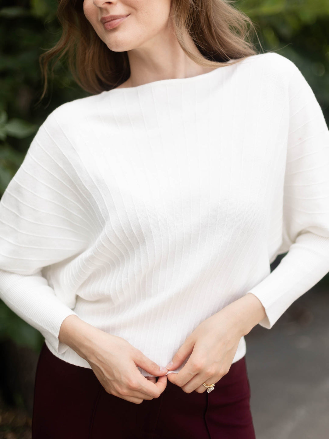 A woman wearing a white ribbed knit long-sleeve top with a boat neckline, paired with dark burgundy pants, shown in an outdoor setting with blurred green foliage in the background.