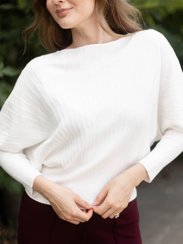 A woman wearing a white ribbed knit long-sleeve top with a boat neckline, paired with dark burgundy pants, shown in an outdoor setting with blurred green foliage in the background.