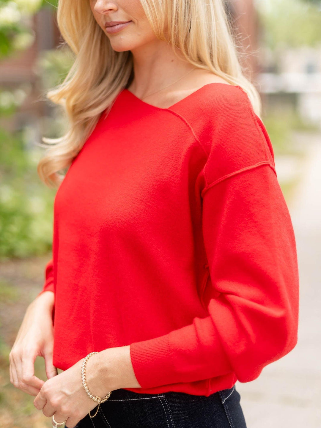 A woman wearing a bright red long-sleeve knit top with a wide boat neckline, shown in an outdoor setting with a blurred background.