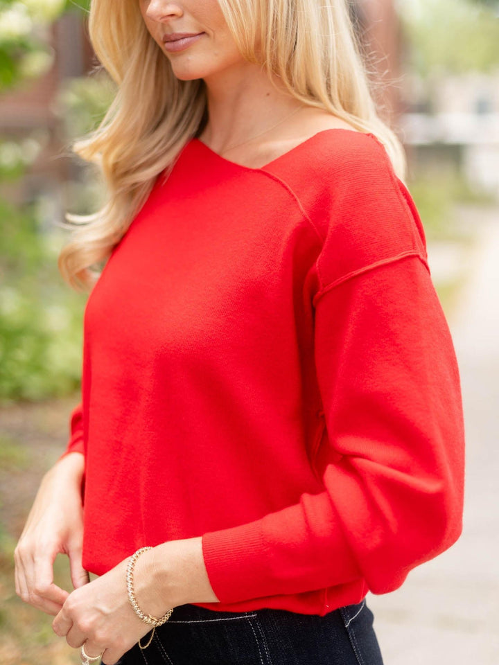 A woman wearing a bright red long-sleeve knit top with a wide boat neckline, shown in an outdoor setting with a blurred background.