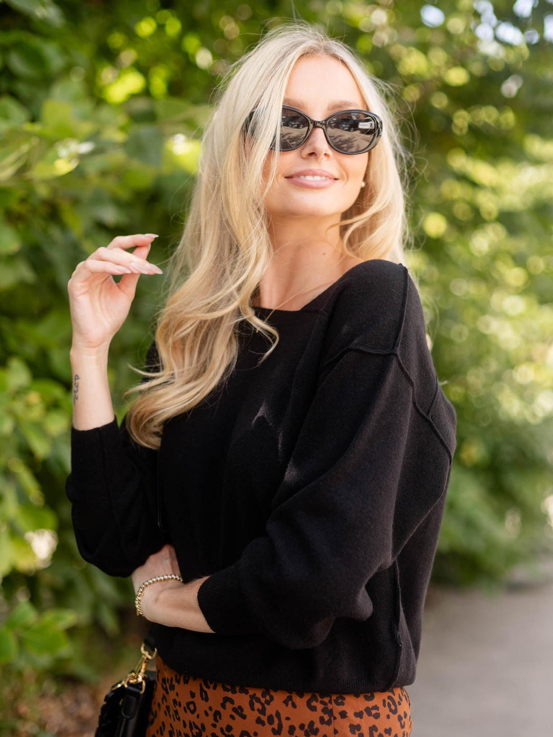 A blonde woman wearing a black knit long-sleeve top and a leopard-print skirt, posing outdoors against a blurred green foliage background.
