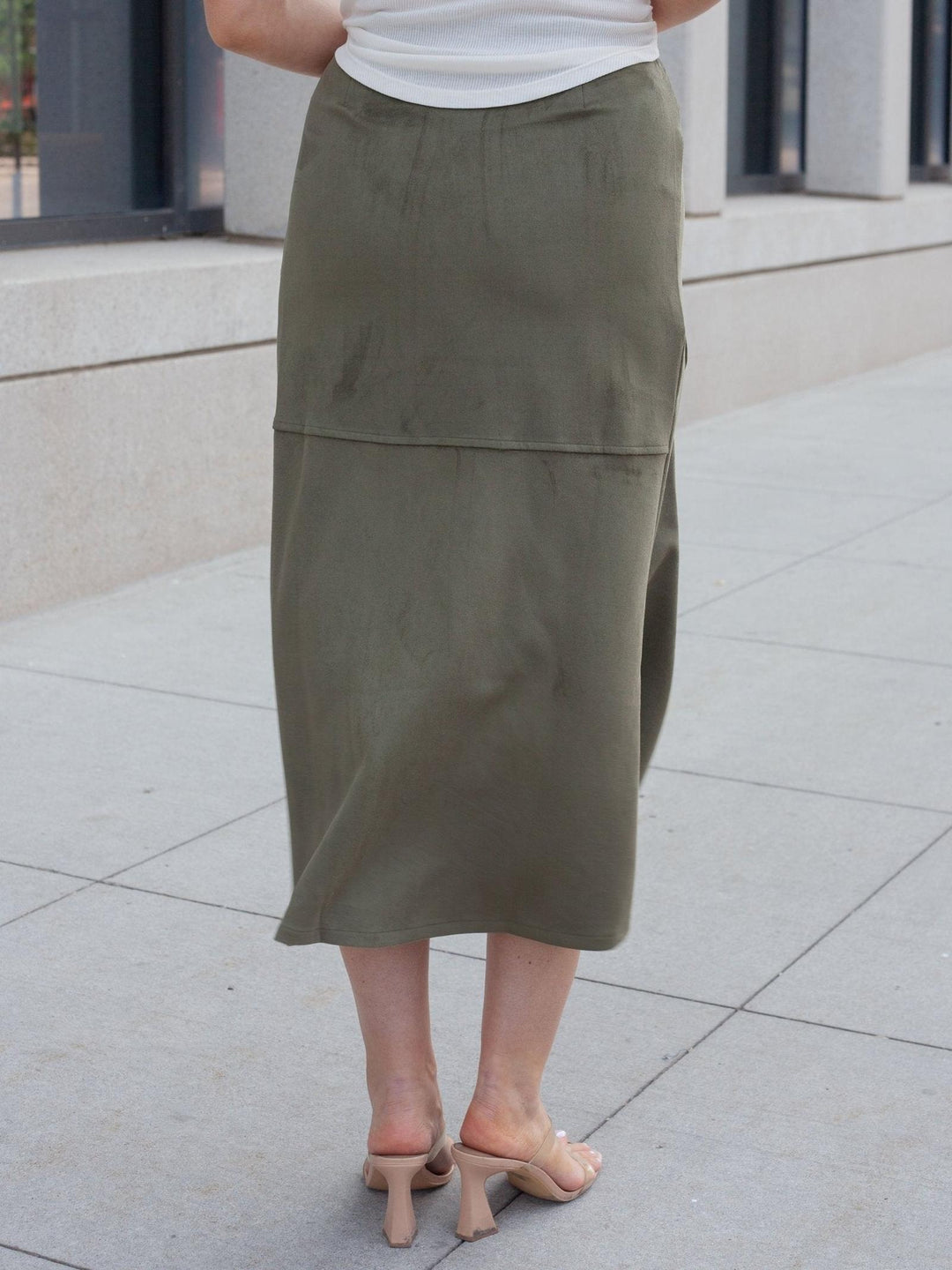 An olive green midi skirt worn by a model, shown from the back in an outdoor urban setting with a concrete sidewalk and building facade in the background.