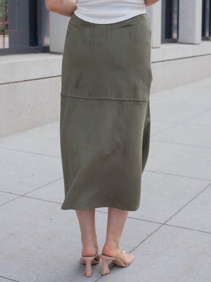 An olive green midi skirt worn by a model, shown from the back in an outdoor urban setting with a concrete sidewalk and building facade in the background.