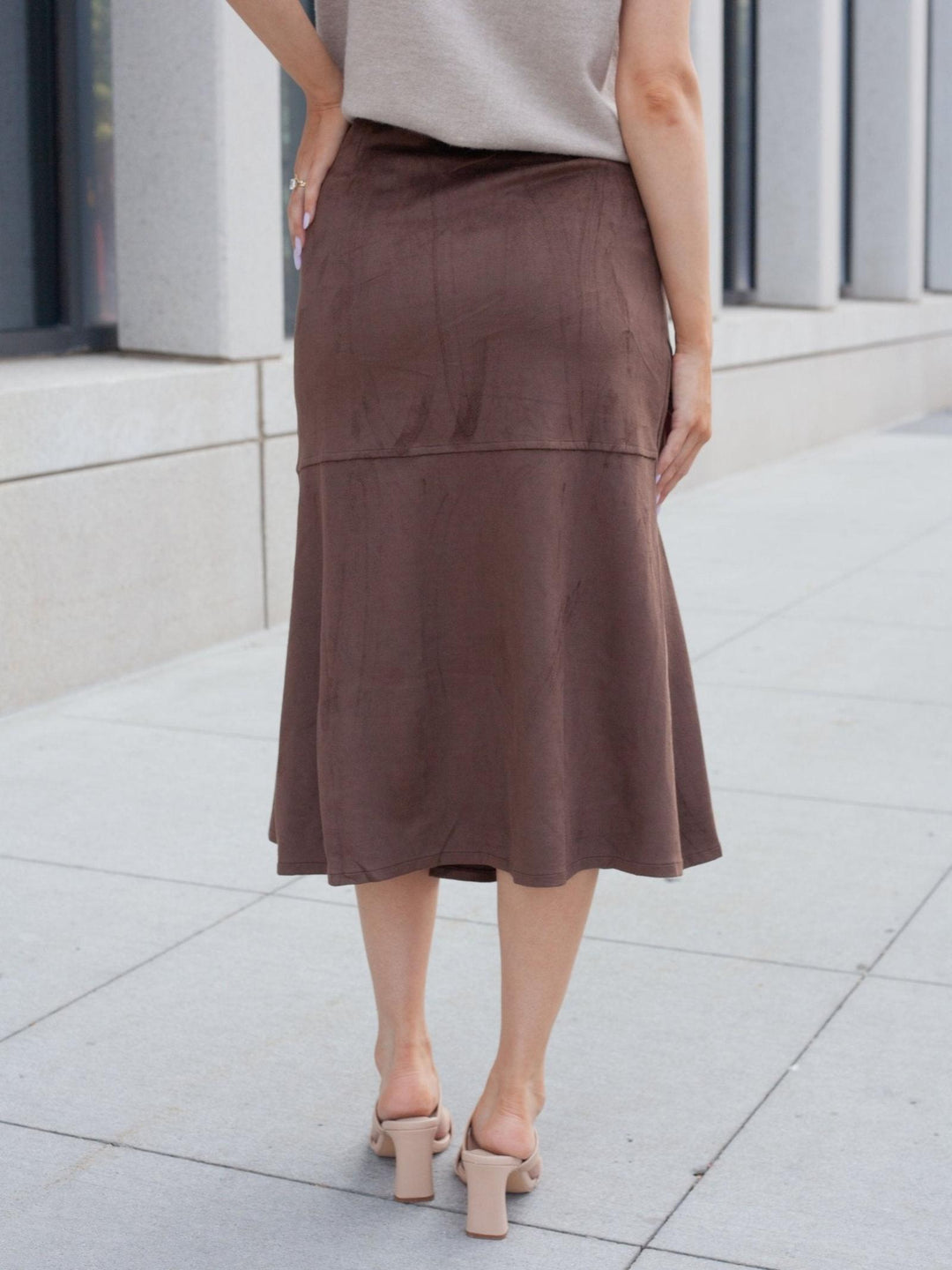 A woman wearing a brown midi skirt with a subtle sheen, paired with a light beige top and beige block-heel mules, standing on a paved outdoor walkway.