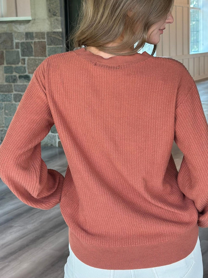 A woman wearing a textured, ribbed knit sweater in a dusty rose color, shown from the back in an indoor setting with a stone wall and wood floor.