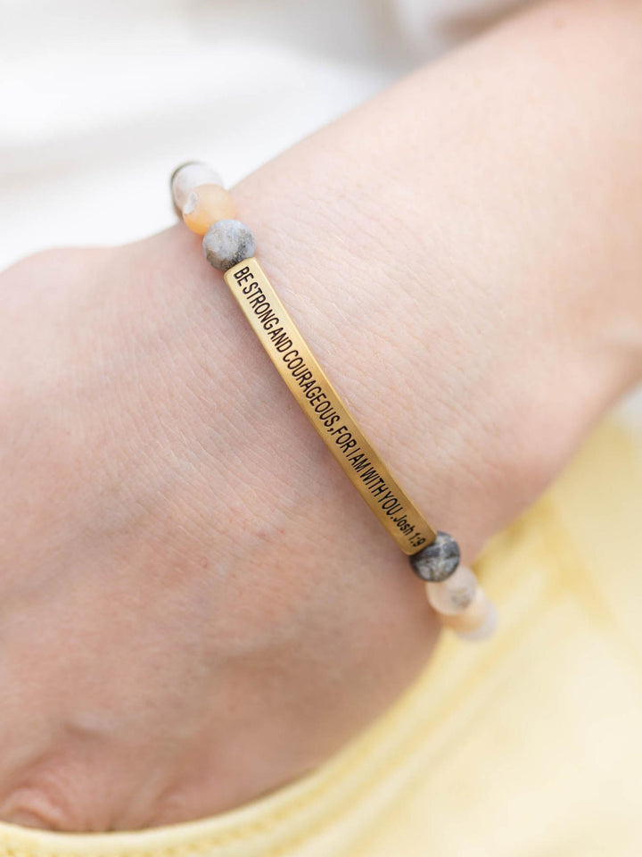 Hand wearing a "Be Strong and Courageous" stone bracelet with a gold bar and light pink/grey beads.