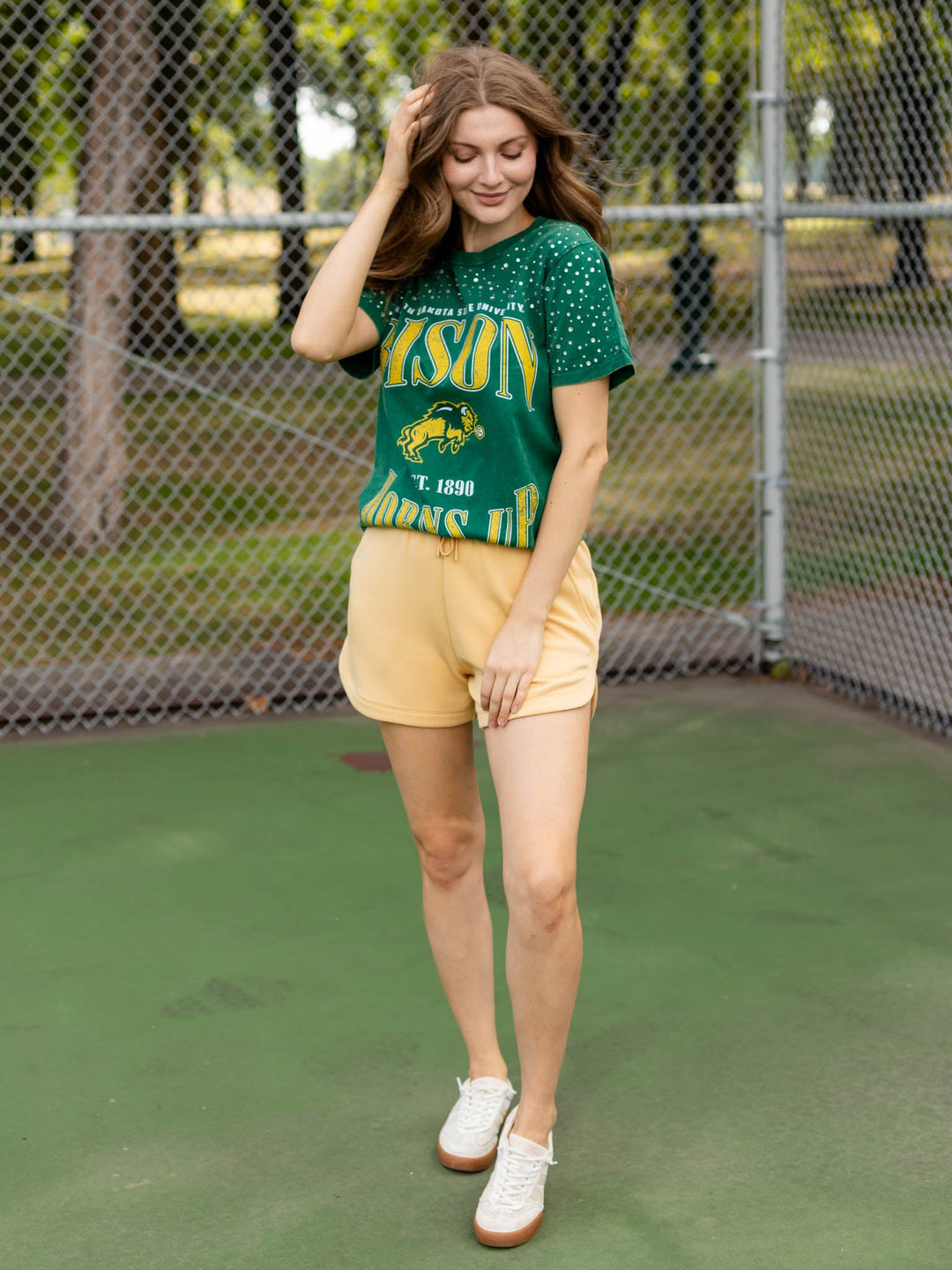 A woman stands on a green outdoor court in front of a chain-link fence, wearing a green and yellow &