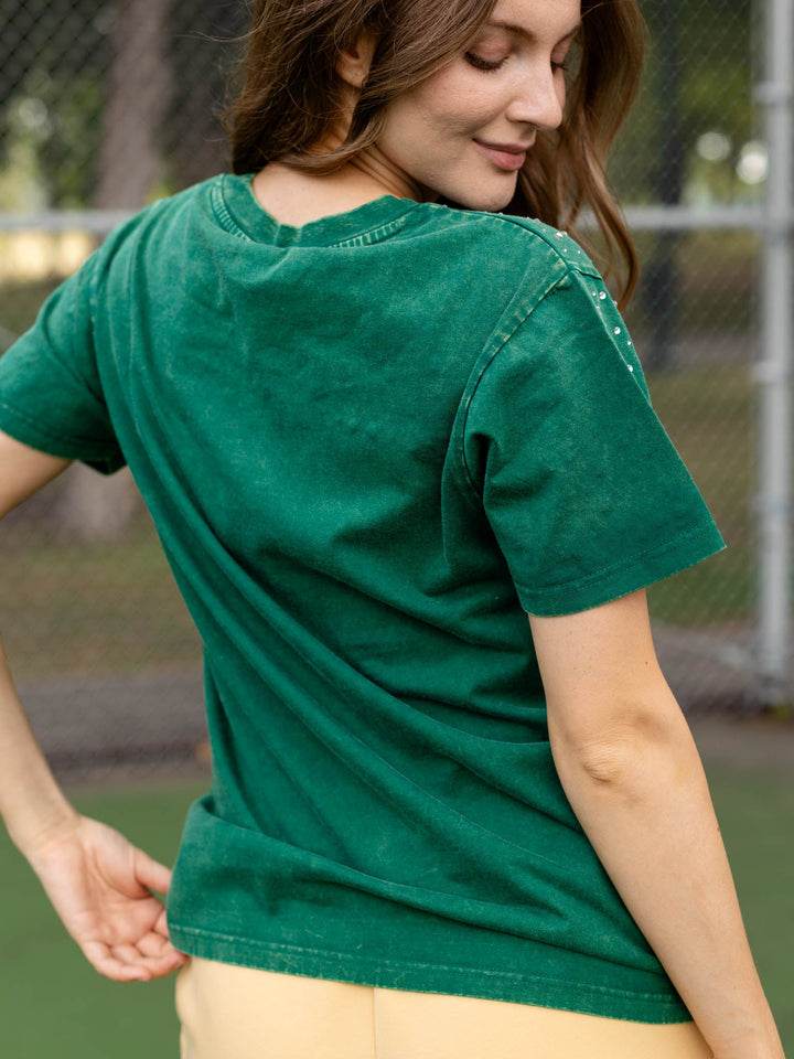 A woman wearing a green short-sleeve t-shirt with a faded, washed texture, shown from a back-three-quarter angle in an outdoor setting with a chain-link fence in the background.