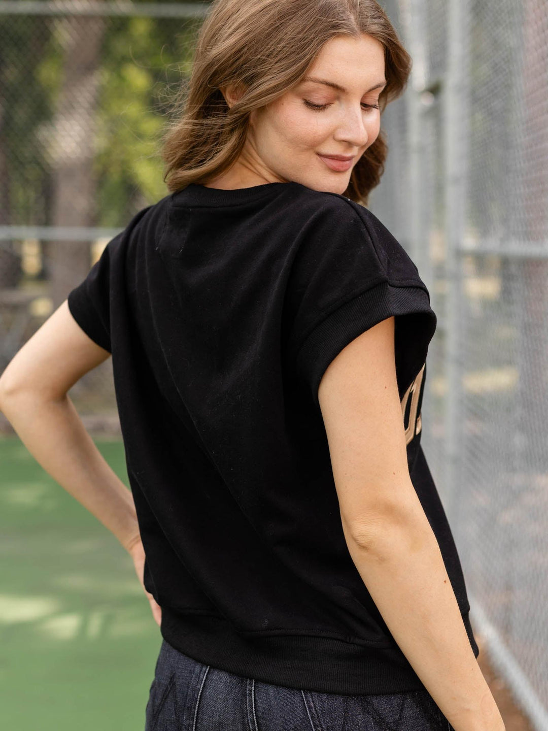 A woman wearing a black short-sleeve t-shirt with a beige graphic print on the back, standing on an outdoor tennis court.