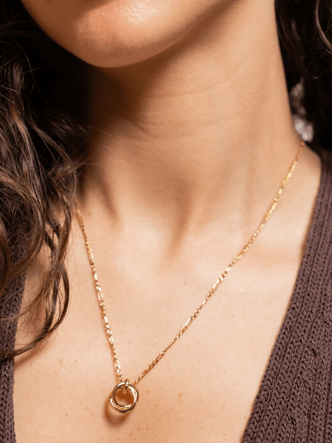 A close-up of a model wearing a delicate gold chain necklace with a small circular pendant featuring a warm amber-orange center, styled with a brown knit top.