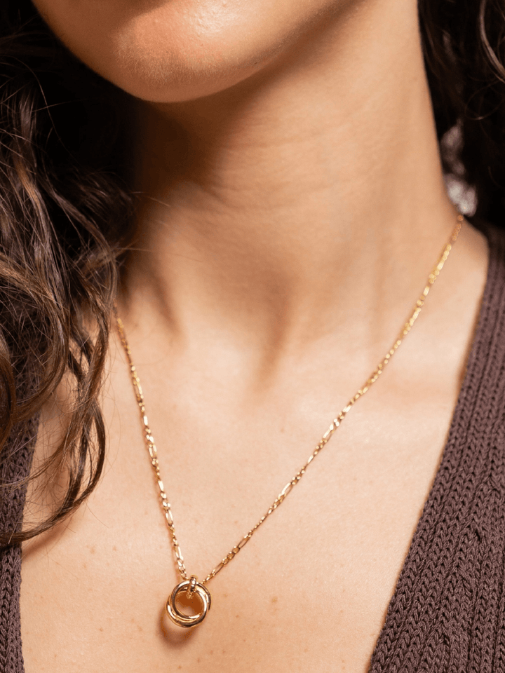 A close-up of a model wearing a delicate gold chain necklace with a small circular pendant featuring a warm amber-orange center, styled with a brown knit top.