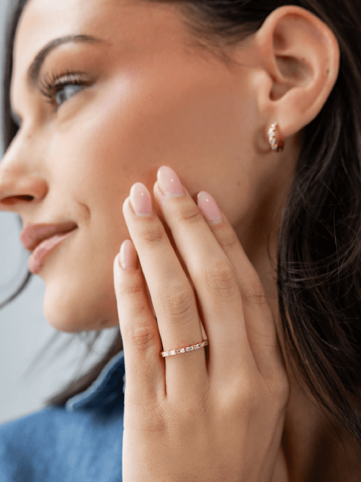A close-up lifestyle shot of a woman wearing a delicate gold band ring with small clear stones on her ring finger, alongside a matching gold hoop earring.