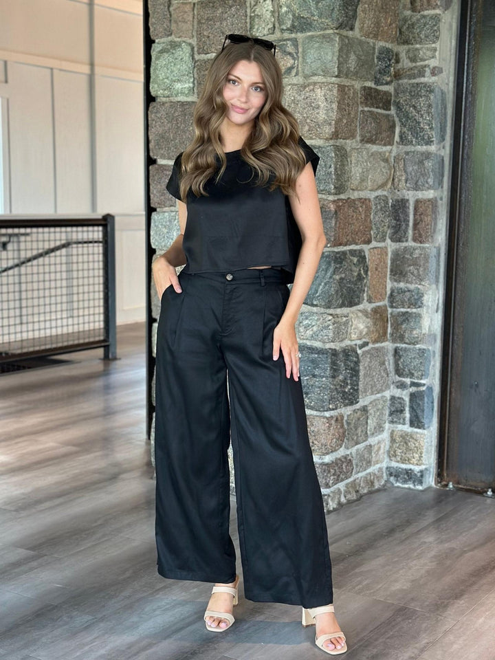 A woman modeling a black sleeveless top and matching black wide-leg pants, standing indoors against a stone wall and wood floor.