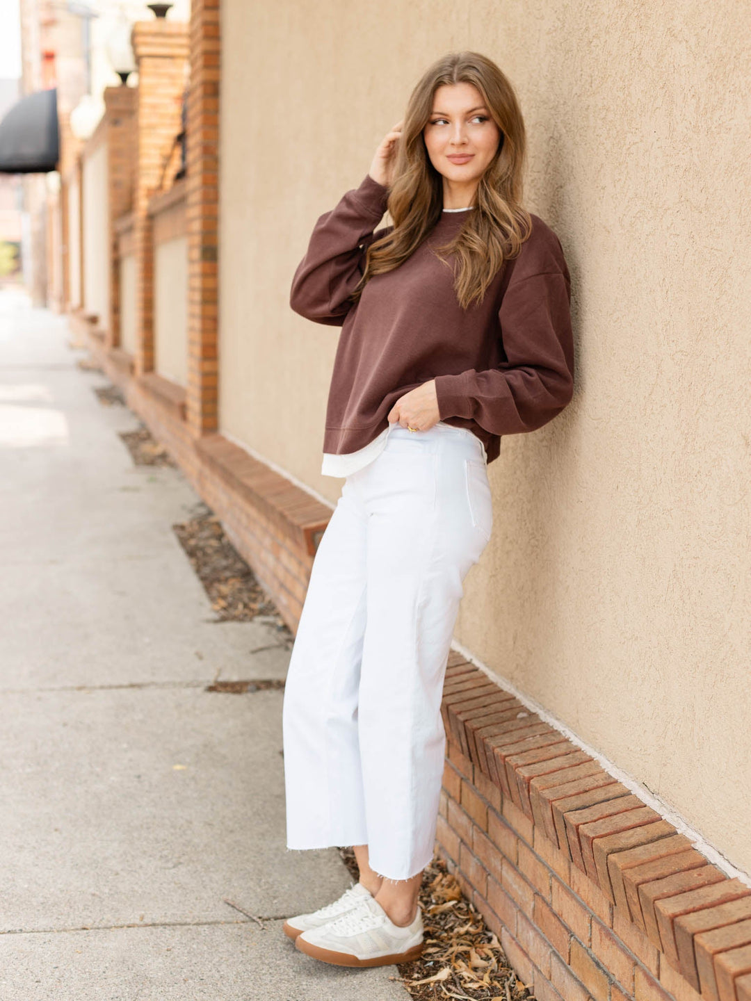 A woman stands on a sidewalk leaning against a beige stucco wall with a brick base. She is wearing a dark brown long-sleeve crewneck sweatshirt, white cropped wide-leg jeans, and white sneakers.