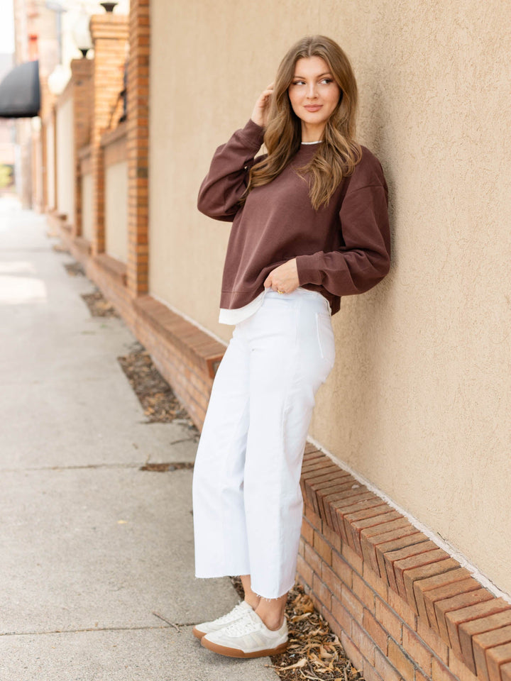 A woman stands on a sidewalk leaning against a beige stucco wall with a brick base. She is wearing a dark brown long-sleeve crewneck sweatshirt, white cropped wide-leg jeans, and white sneakers.