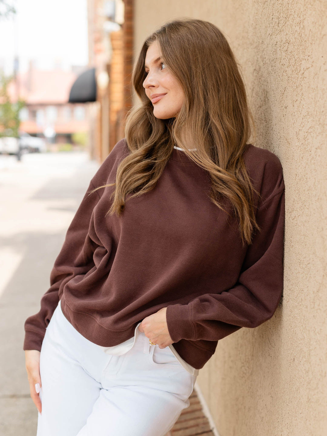 A woman with long brown hair leans against a beige stucco wall, wearing a dark brown crewneck sweatshirt and white pants.