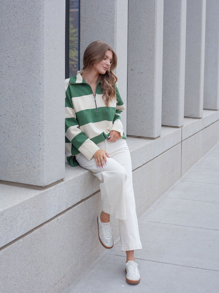 A woman sits on a concrete ledge wearing a green and cream striped half-zip pullover, white wide-leg cropped pants, and white sneakers with brown soles, set against a modern concrete column background.