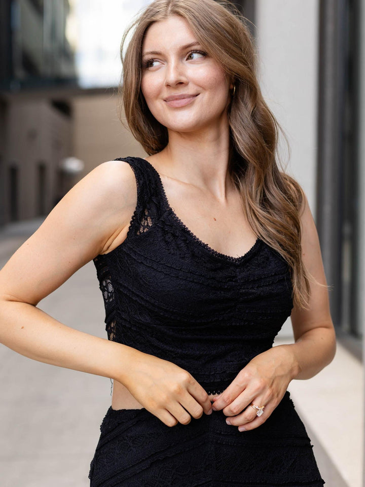 A woman wearing a black lace sleeveless top and matching black skirt, posing in an outdoor urban setting.