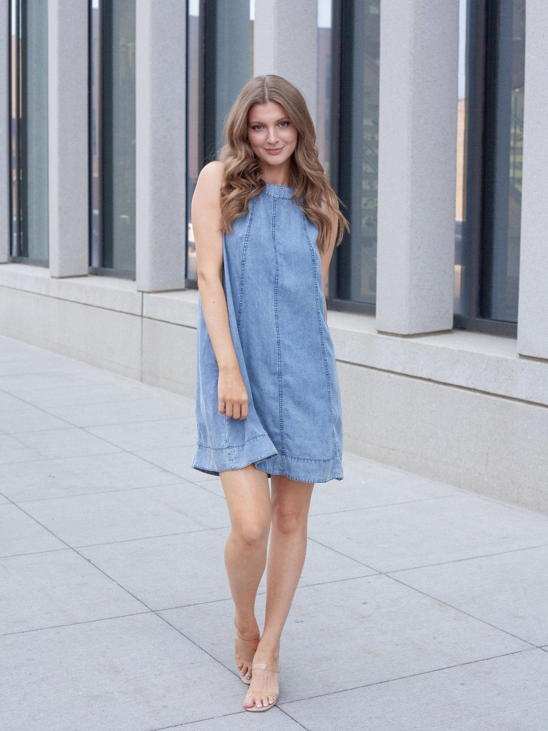 A woman wearing a sleeveless light blue denim dress walking outdoors on a paved walkway in front of a modern building with large glass windows.