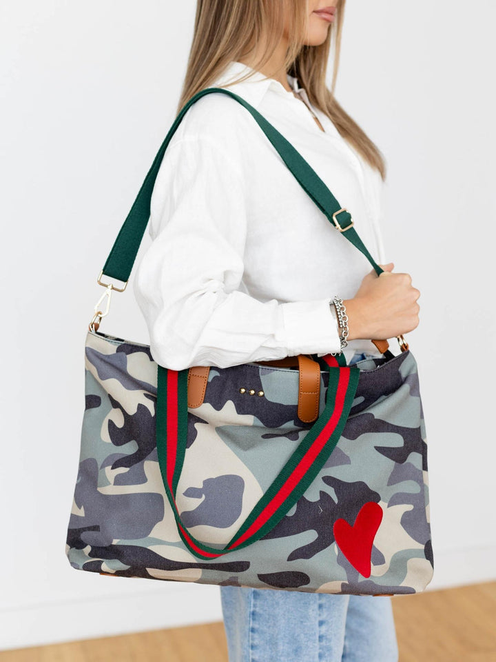 Person holding a camouflage tote bag with green straps and a red heart on a white background