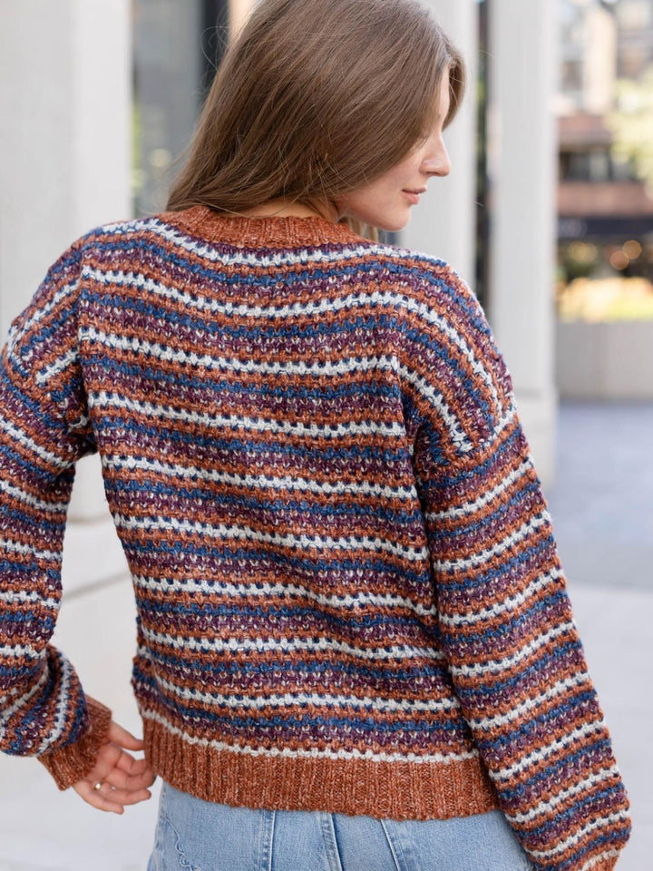 A woman wearing a chunky knit sweater with horizontal stripes in rust orange, navy blue, and off-white, shown from the back in an outdoor setting.