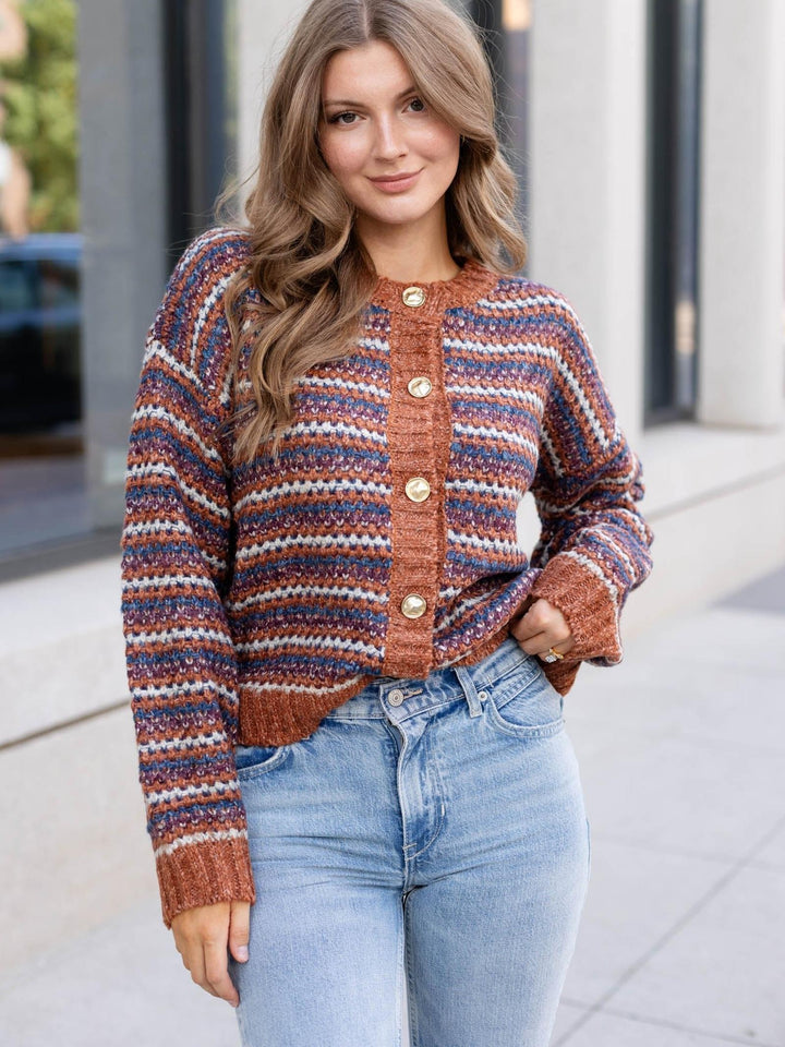A woman wearing a rust-colored knit cardigan with horizontal stripes in blue, beige, and brown, paired with light-wash blue jeans, standing outdoors on a sidewalk.