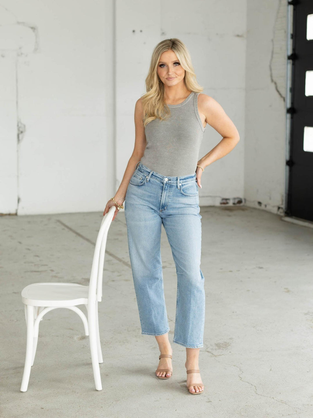 Woman wearing a gray tank top and light blue jeans standing next to a white chair in an indoor setting.